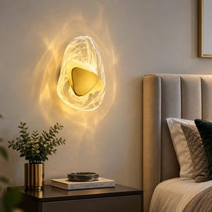 Pode incluir: Um candeeiro de parede com um abajur de vidro translúcido de forma irregular e um padrão branco em espiral. Um elemento central de metal dourado em forma triangular está posicionado no interior, emitindo uma luz quente e suave.