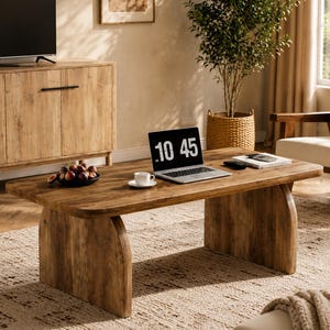May include: A rectangular wooden coffee table with a smooth, light brown wood grain finish and two curved, solid panel legs. The table surface holds a laptop displaying the time 10:45, a small white ceramic cup on a saucer, and a bowl of dark fruit.