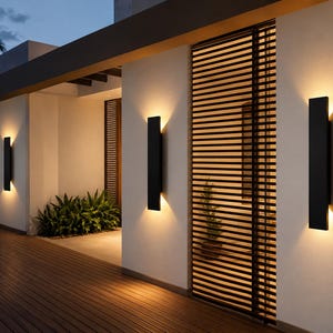 May include: Three rectangular black wall sconces mounted on a white exterior wall. Each fixture emits a warm, vertical glow of light from both the top and bottom, creating a bright illumination pattern against the smooth surface of the building.