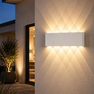 Peut inclure: Une applique murale rectangulaire blanche fixée sur un mur extérieur beige. Le luminaire émet une lumière chaude formant un motif en losange distinct, projetant des faisceaux vers le haut et vers le bas sur la surface plane du mur.