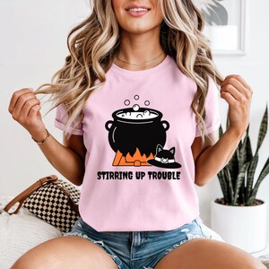Op de afbeelding: Een lichtroze t-shirt met een afbeelding van een zwarte ketel boven oranje vlammen met witte bubbels en een zwarte hoed met kattenoren. De tekst STIRRING UP TROUBLE staat in zwarte hoofdletters onder de afbeelding van de ketel.