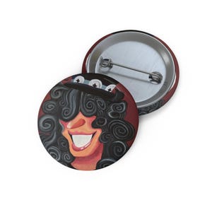 May include: A round pin-back button featuring a stylized illustration of a person with voluminous dark curly hair wearing a black top hat with white star accents. The person has a wide, smiling mouth and a red-toned background. The back is silver metal.