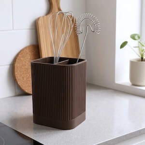 Può includere: Un portautensili rettangolare marrone scuro con una texture a coste verticali sulle pareti esterne. Il contenitore è dotato di un divisorio centrale e contiene due fruste da cucina in metallo argentato di diverso design.