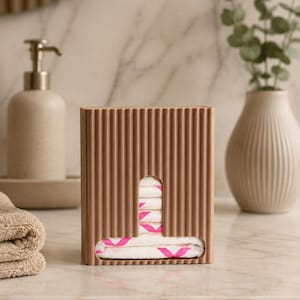 May include: A rectangular, ribbed, brown plastic dispenser box featuring a T-shaped cutout on the front. Inside the dispenser, a stack of white sanitary pads with a bright pink graphic pattern is visible through the opening.