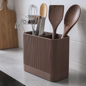 Op de afbeelding: Een rechthoekige bruine keukengereedschaphouder met een verticale geribbelde textuur en een verdeelde binnenkant. De houder bevat een metalen garde, een metalen tang, een houten spatel en een houten lepel, die allemaal rechtop staan.