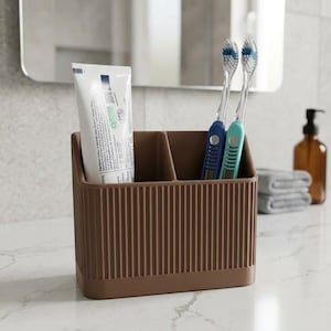 May include: A brown rectangular bathroom organizer with vertical ribbed detailing and two separate compartments. One compartment holds a white tube of toothpaste, while the other holds two toothbrushes with blue and light blue handles.