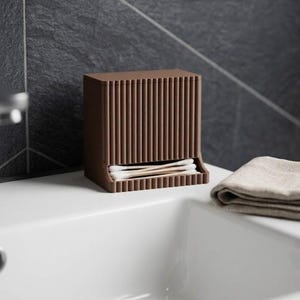 May include: A brown, rectangular cotton bud dispenser featuring a vertical ribbed texture on its exterior. The container has an open front slot at the base where several white cotton buds with brown paper stems are visible and accessible.