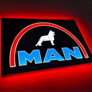 Può includere: Un'insegna da parete rettangolare che presenta il logo MAN con una silhouette di leone bianca sopra il testo blu MAN, racchiuso da un bordo ad arco rosso. L'insegna è retroilluminata, proiettando un vivido bagliore rosso sulla parete.