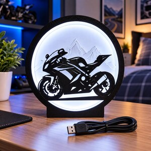 Può includere: Una lampada LED circolare nera che presenta la silhouette di una moto sportiva su uno sfondo di catena montuosa retroilluminato in bianco. La lampada ha una base piatta e include un cavo di alimentazione USB nero con connettore standard.