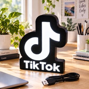 Può includere: Un'insegna luminosa nera con il logo TikTok, caratterizzata dall'icona della nota musicale bianca e dalla scritta TikTok in lettere bianche. L'insegna ha una cornice nera e include un cavo di alimentazione USB nero con connettore standard.
