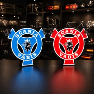 May include: Two circular Scania Vabis illuminated signs, one with a blue background and one with a red background. Both signs feature the black griffin logo with a crown and the text SCANIA VABIS in white. Each sign includes two clear suction cups.