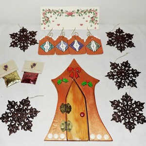 May include: A set of Christmas ornaments featuring a wooden door with a red bow, holly, and gold hinges. The set also includes six brown glitter snowflakes and two small bags of glitter.