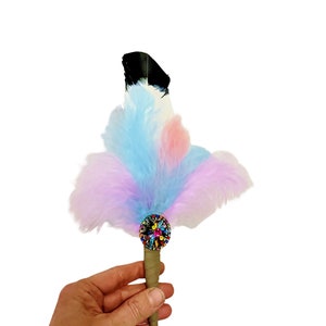 May include: A colorful feather pen with a multi-colored jeweled embellishment at the base of the feather. The pen is held in a hand.