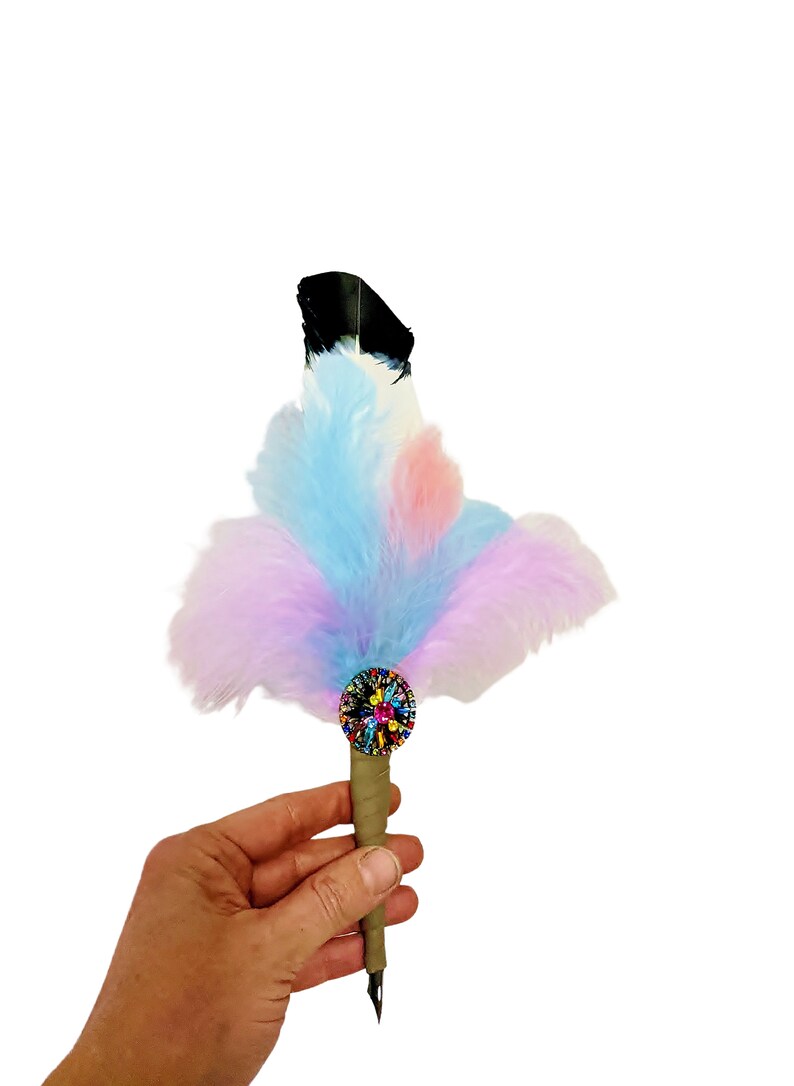 May include: A colorful feather pen with a multi-colored jeweled embellishment at the base of the feather. The pen is held in a hand.