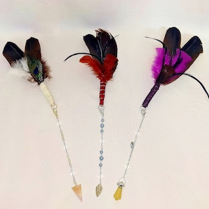 May include: Three feather wands with different colored feathers and crystals on the end of a chain. The wands are made with a wooden handle and have a variety of feathers including peacock, black, red, and pink.