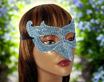 May include: A light blue masquerade mask with intricate lace-like detailing and sparkling accents. The mask features a pointed nose and is secured with a black elastic band. The mask is worn by a mannequin with brown hair and red lipstick.