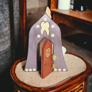 May include: A purple wooden house with a small red door and a white tooth design. The house is sitting on a brown wooden surface.