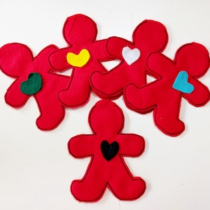 May include: Five red felt gingerbread man ornaments with black stitched outlines. Each ornament has a heart in a different color: green, yellow, white, and blue. The fifth ornament has a black heart.
