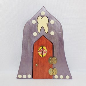 May include: A purple wooden fairy door with a tooth design above the door. The door has a small stained glass window and brass hinges.