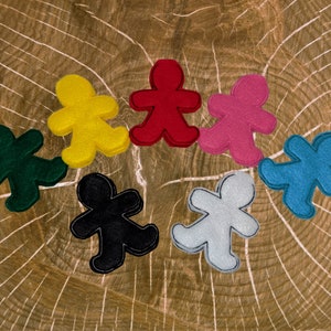 May include: Seven felt figures of people in different colors, including red, yellow, green, blue, pink, black, and white. The figures are arranged in a circle on a wooden surface.