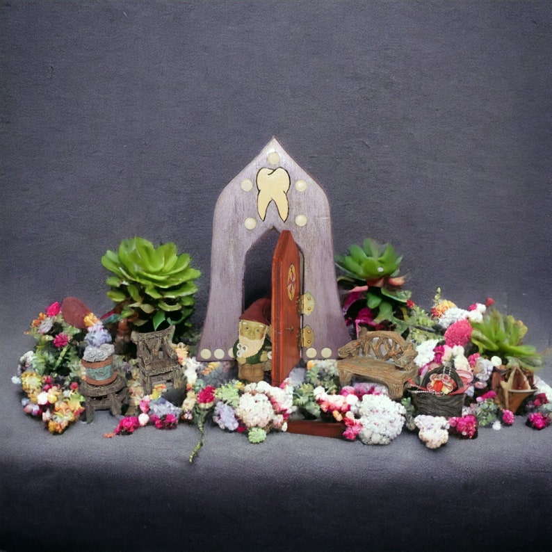 May include: A miniature wooden fairy house with a purple door and a tooth-shaped design. The house is surrounded by small plants and flowers, including succulents and a gnome figurine.