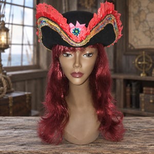 May include: A black tricorn hat featuring a vibrant red lace trim, a decorative gold and multicolored sequin border, and a central pink fabric flower with a blue gemstone accent, displayed on a mannequin head with long wavy red hair.