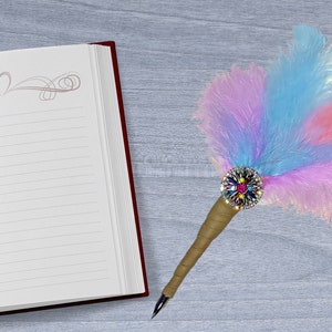 May include: A blank lined journal with a red cover and a decorative heart design on the first page. A feather pen with a colorful feather and a jeweled embellishment is lying on a gray surface next to the journal.