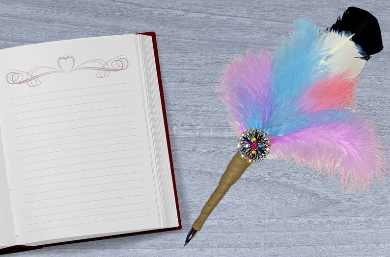 May include: A blank lined journal with a red cover and a decorative heart design on the first page. A feather pen with a colorful feather and a jeweled embellishment is lying on a gray surface next to the journal.