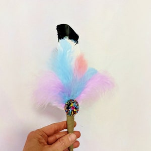May include: A colorful feather pen with a black tip and a multi-colored jeweled embellishment. The pen is held in a hand against a white background.