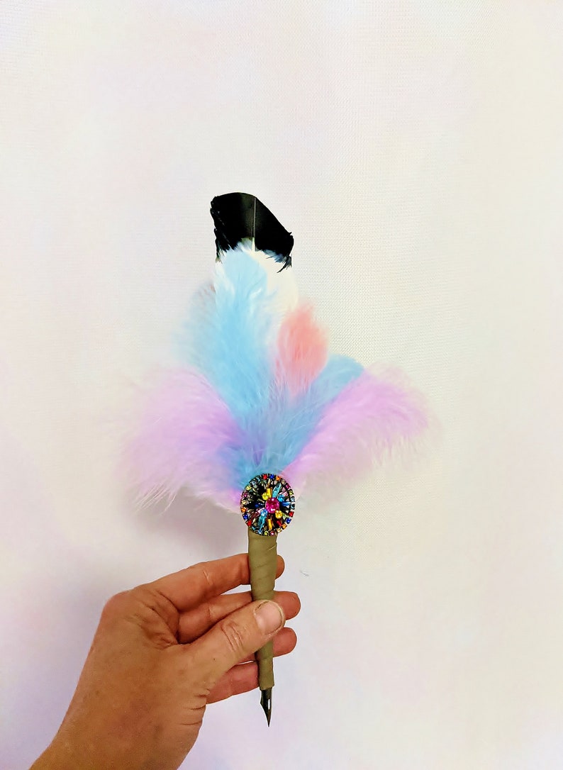 May include: A colorful feather pen with a black tip and a multi-colored jeweled embellishment. The pen is held in a hand against a white background.