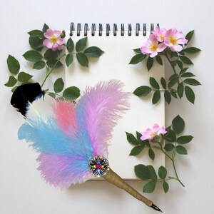 May include: A blank notebook with a spiral binding surrounded by pink rose blossoms and a colorful feather pen with a jeweled tip.