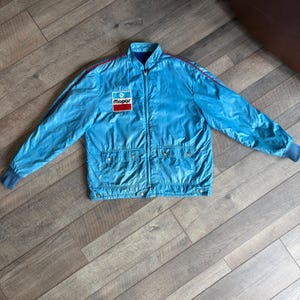 May include: A light blue zip-up jacket featuring a Mopar patch on the chest, red piping along the shoulders and sleeves, two front pockets with snap closures, and ribbed cuffs. The jacket has a high collar and a smooth, lightweight finish.