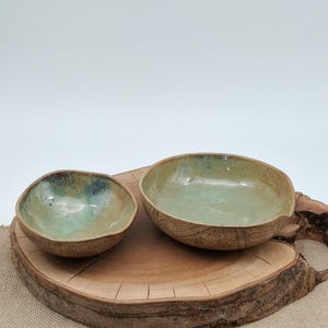 Può includere: Due ciotole in ceramica fatte a mano con interno smaltato verde chiaro ed esterno marrone testurizzato con intricati motivi a linee. Le ciotole hanno forme organiche irregolari e sono presentate insieme come un set.