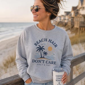 Könnte beinhalten: Ein graumeliertes Sweatshirt mit Rundhalsausschnitt und einem Grafikdruck mit dem Text BEACH HAIR DON'T CARE SALT & SKY, Palmen und einer Sonne. Ein weißer Isolierbecher mit dem Text COFFEE COAST REPEAT und einem Anker-Symbol ist ebenfalls zu sehen.
