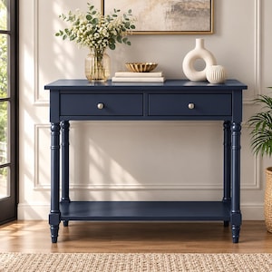 Puede incluir: Una mesa consola de color azul marino con dos cajones con pomos plateados, patas torneadas y un estante inferior. La mesa rectangular tiene un acabado liso y una estructura robusta, proporcionando una superficie plana para el almacenaje.