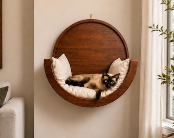 May include: A circular, wall-mounted wooden cat bed with a dark brown finish, featuring a plush, cream-colored cushion lining the interior. A Siamese cat with dark brown points and a tan coat is resting inside the curved wooden structure.