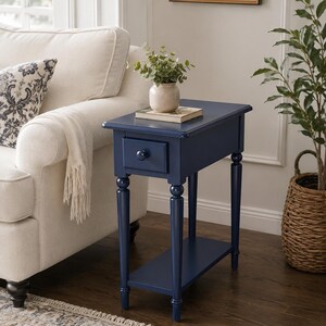 Puede incluir: Una mesa auxiliar estrecha y rectangular de color azul marino con un cajón con un pequeño pomo redondo, cuatro patas torneadas delgadas y un estante inferior. La superficie sostiene un jarrón beige con follaje verde y un libro.