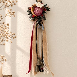 May include: A wall hanging decoration featuring a cluster of dried flowers, a dried orange slice, star anise, and red berries. Below the floral arrangement hang several ribbons in burgundy, black, and cream lace, along with three small metal bells.