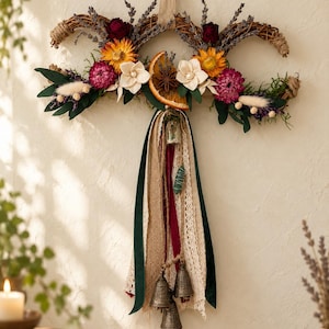 May include: A crescent-shaped wall hanging made of woven twigs, decorated with dried lavender, yellow sunflowers, dark red roses, white wood flowers, and a dried orange slice. Long ribbons in green, cream lace, and burlap hang with three metal bells.