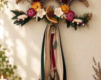 May include: A crescent-shaped wall hanging made of woven twigs, decorated with dried lavender, yellow sunflowers, dark red roses, white wood flowers, and a dried orange slice. Long ribbons in green, cream lace, and burlap hang with three metal bells.
