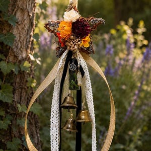 May include: A hanging star-shaped ornament crafted from woven twigs, adorned with dried flowers in shades of orange, cream, and deep red. It features three brass bells, a small pentagram charm, a clear glass vial, and long ribbons.