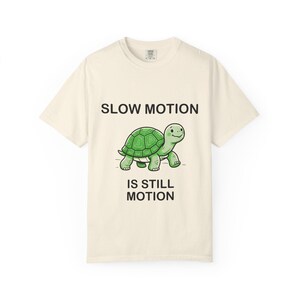 Peut inclure: Un t-shirt à manches courtes de couleur crème avec un motif central représentant une tortue verte. Au-dessus de la tortue, le texte indique SLOW MOTION, et en dessous, le texte indique IS STILL MOTION. Le motif est imprimé en noir.
