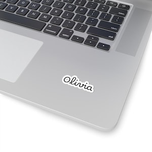 Peut inclure: Un autocollant blanc découpé à la forme présentant le prénom Olivia écrit dans une police noire, arrondie et cursive. L'autocollant est appliqué sur la surface d'un ordinateur portable argenté, sous la zone du clavier près du pavé tactile.