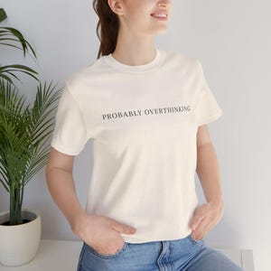 Peut inclure: Un t-shirt à manches courtes de couleur crème avec le texte noir "PROBABLY OVERTHINKING" imprimé sur la poitrine. Le t-shirt présente une encolure ronde classique et un ourlet droit, associé à un jean bleu, pour un style décontracté.