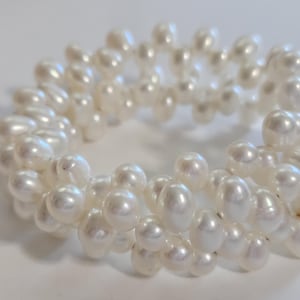 May include: A multi-strand bracelet composed of numerous small, lustrous white freshwater pearls. The pearls are arranged in a dense, clustered circular formation, creating a textured, layered appearance with a reflective, smooth surface finish.