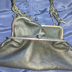 May include: A dark olive green leather handbag featuring a braided shoulder strap and a silver-toned metal kiss-lock closure. A smaller matching coin purse with a kiss-lock frame is attached to the main bag, accented with colourful stitching.