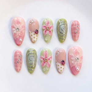May include: A set of ten almond-shaped press-on nails featuring a variety of designs including 3D pink flowers with rhinestones, green and white polka dots with ladybug accents, silver swirls on sage green, and pink and white plaid patterns.