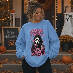 May include: A light blue crewneck sweatshirt featuring a graphic print of a masked figure in a black hooded robe holding a pink rotary telephone. The text reads "SCREAM QUEEN" and "DON'T HANG UP" in pink, surrounded by pink hearts and roses.