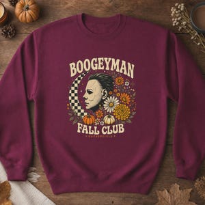 May include: A maroon crewneck sweatshirt featuring a graphic print with the text BOOGEYMAN FALL CLUB and HADDONFIELD. The design includes a profile of a masked figure, a checkered crescent moon, various autumn flowers, and two small pumpkins.