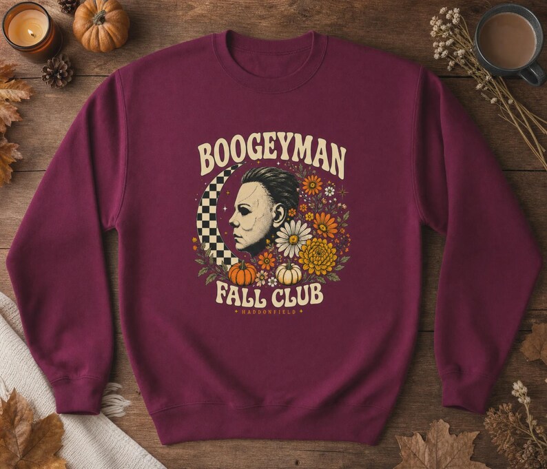 May include: A maroon crewneck sweatshirt featuring a graphic print with the text BOOGEYMAN FALL CLUB and HADDONFIELD. The design includes a profile of a masked figure, a checkered crescent moon, various autumn flowers, and two small pumpkins.